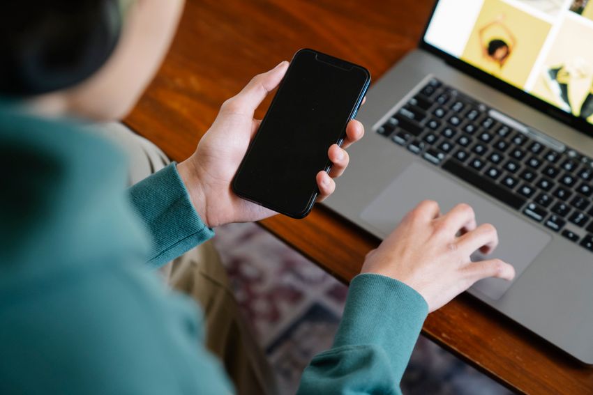 Eine Person hält ein Smartphone und bedient daneben einen Laptop auf einem Holztisch